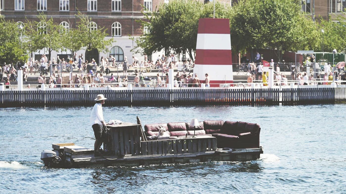 A man steers a large sofa on a raft along a river with crowd 1cd8e933e84c11bddabb68c436c52f93