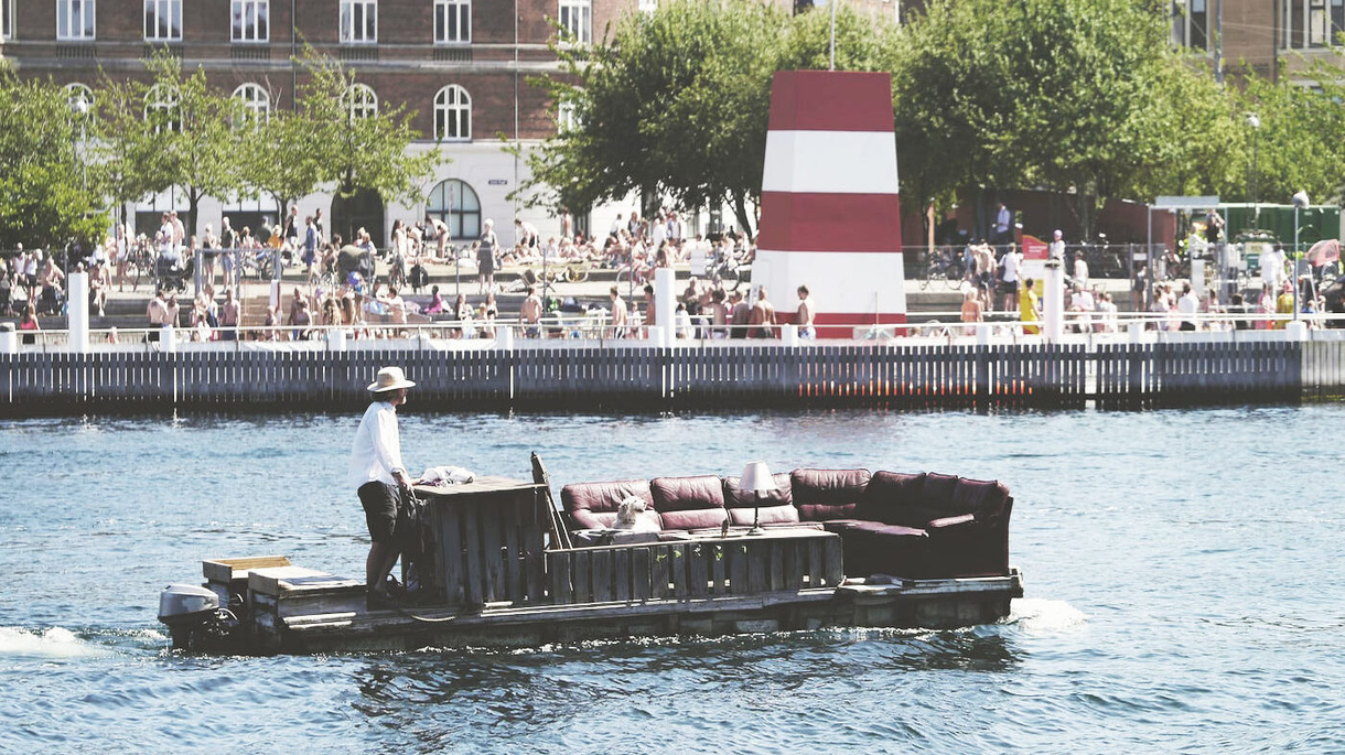 A man steers a large sofa on a raft along a river with crowd 1cd8e933e84c11bddabb68c436c52f93