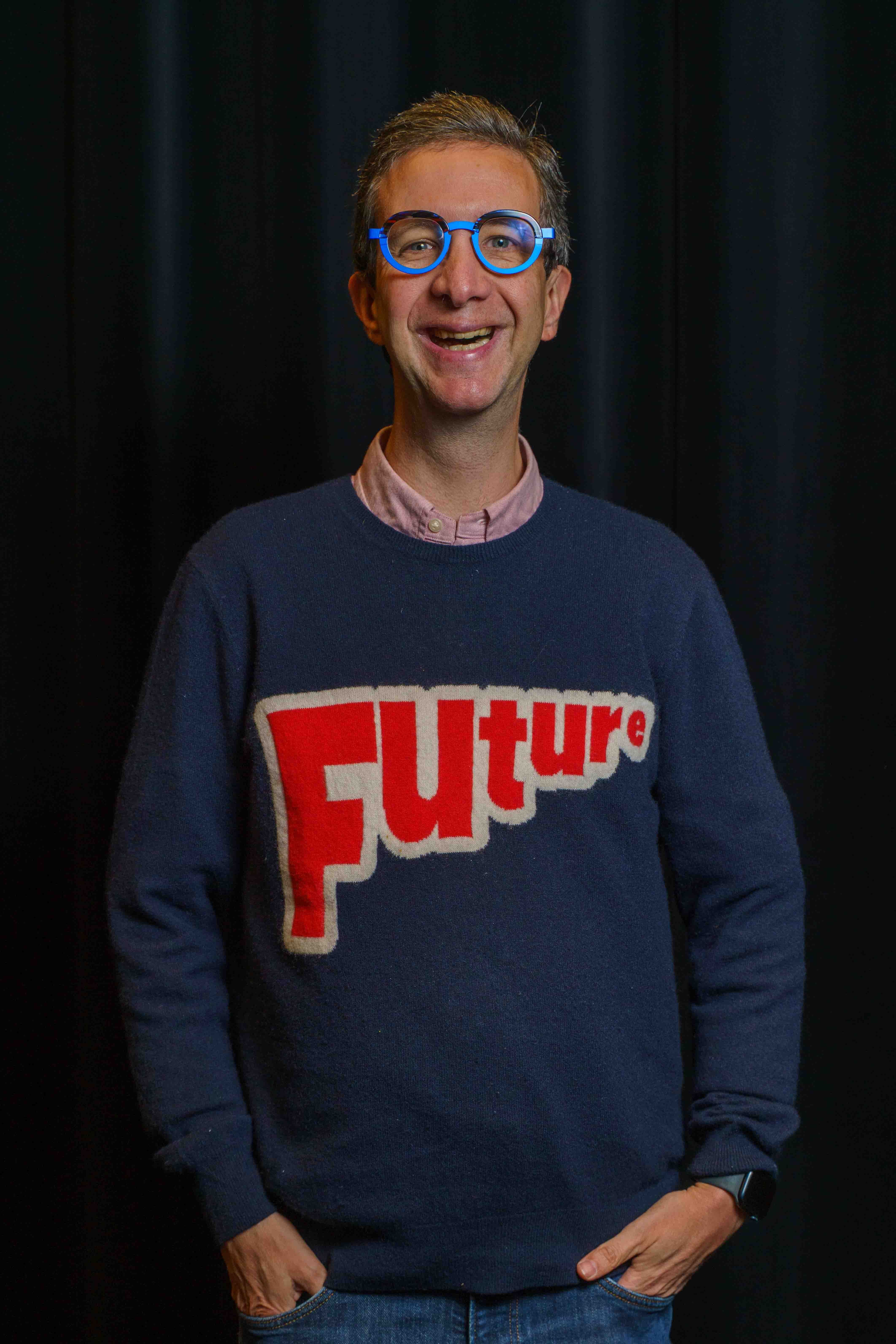 Portret van Nick voor een zwart toneelgordijn. Hij heeft een ronde blauwe bril op en lacht naar de camera. Op zijn blauwe trui staat in dikke rode letters 'Future'.