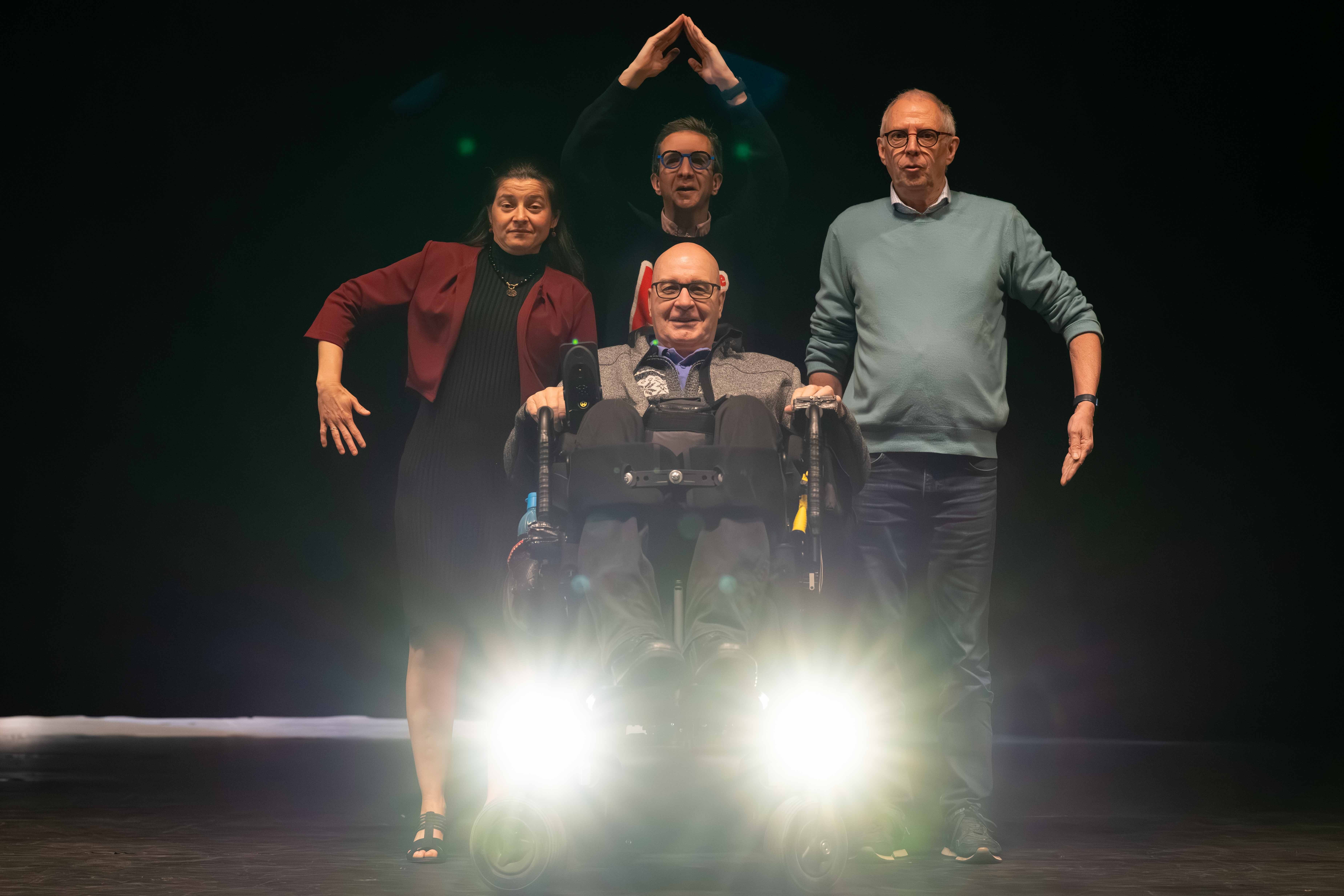 Vier mensen staan op het podium voor een zwart theaterdoek. Vooraan zit een persoon in een rolstoel met twee koplampen, links en rechts flankeren twee mensen hem, achter hem staat iemand met de armen in de lucht.